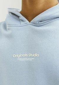 Felpa leggera blu chiaro realizzata in tessuto morbido, con tasca frontale, cappuccio con coulisse e testo del logo bianco: "Originals Studio" sul petto.