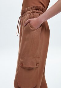 Pantalon léger marron avec taille élastique, cordon de serrage, poche latérale et deux poches cargo boutonnées sur les jambes.