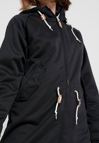Czarna kurtka zapinana na zamek błyskawiczny z kapturem, wykonana z gładkiego materiału. Wyposażona w białe sznurki i brązowe akcenty na suwakach i kieszeniach.