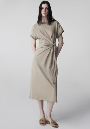 Jeune femme aux longs cheveux foncés portant une robe midi beige avec un noeud sur le côté et des sandales plates ouvertes couleur beige, debout devant un fond uni.