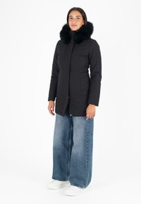 Parka nera con colletto in pelliccia nera, abbinata a jeans a gamba larga blu e sneakers bianche. Silhouette pulita e moderna.