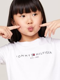 T-shirt di cotone bianco con il logo "TOMMY HILFIGER EST. 1985" in nero e un piccolo accento rosso e blu. Ragazza che si pizzica le guance in modo giocoso.
