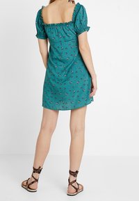 Tyrkysové šaty s čtvercovým výstřihem, nadýchanými rukávy a květinovým vzorem. V kombinaci s hnědými sandály na pásky s texturovaným povrchem.