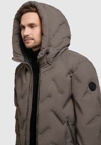 Uomo che indossa una giacca a vento color taupe con cappuccio alzato, zip a metà e mani fuori dal fotogramma, su uno sfondo grigio uniforme.