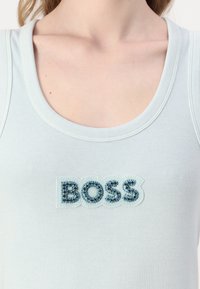 Svjetloplavi rebrasti top bez rukava s teksturiranim aplikama "BOSS" ukrašenim plavim rhinestones na prednjoj strani.