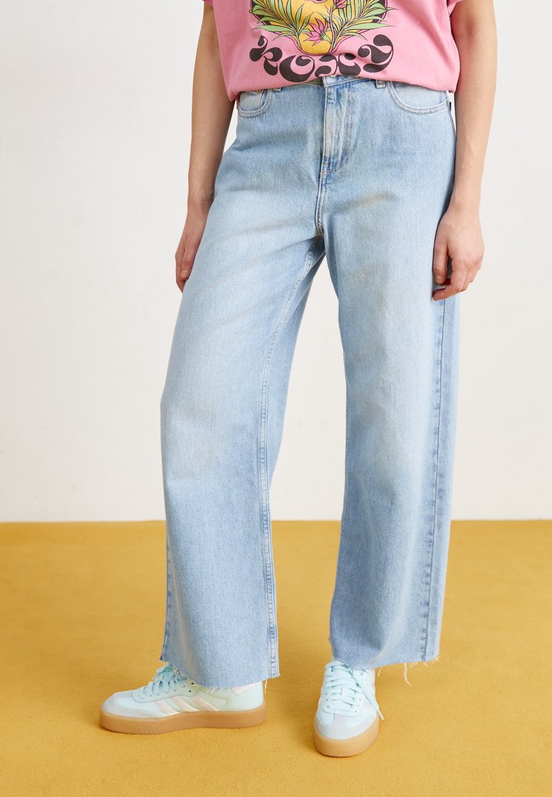 Vaalean siniset farkut, joissa on leveä lahje ja korkeavyötäröinen malli, hemmojen reunoilla on hieno rispaantuminen. Yhdistetty mintunvihreisiin tennareihin.