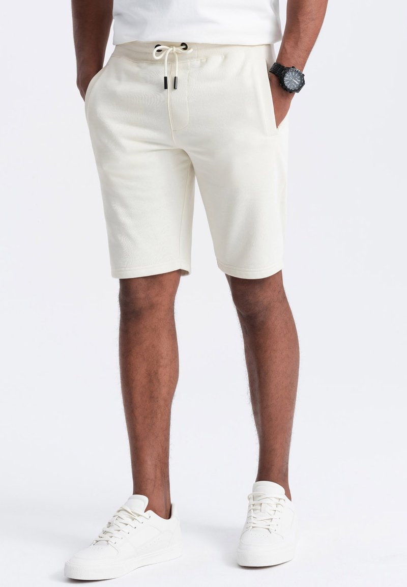 Pantalones cortos de color crema con cintura elástica y cordón. Textura suave, corte a la altura de la rodilla, combinados con zapatillas blancas.