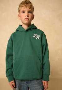 Hoodie vert en tissu épais, avec une poche avant et une broderie blanche « LA » sur la poitrine, associé à un jean bleu.