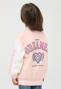 Giubbotto bomber rosa con maniche bianche, con testo "LITTLE DREAMER" e grafica di un cuore con ali. Polsini e orlo a coste.