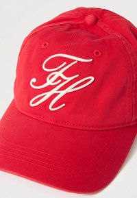 Červená bavlnená čiapka s bielym vyšívaným nápisom na prednej strane, zahnutým šiltom a gombíkom na vrchu. Textúrovaná tkanina s prešívanými detailmi.