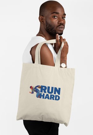 Muž s vousy a hodinkami drží béžovou tašku přes rameno s motivem vintage fotbalisty a modrým textem "RUN HARD" na bílém pozadí.