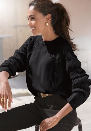 Black knitted sweater with textured patterns on sleeves and shoulders. Paired with high-waisted black trousers and a gold belt.