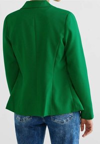 Zielona, dopasowana marynarka o zarysowanej sylwetce, z klapami z wycięciem i gładką fakturą materiału, zestawiona z niebieskimi dżinsami.