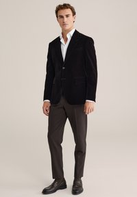 Juodas aksominis švarkas su išpjautomis atlapais, vienu mygtuko užsegimu ir dviem priekinėmis kišenėmis, derinamas su balta marškiniais, rudos spalvos kelnėmis ir juodais batais.