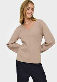 Femme souriante avec des cheveux blonds posant avec un pull beige en V et un pantalon sombre devant un fond clair et uni.