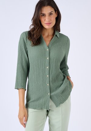 Femme portant un chemisier texturé vert sauge à boutons avec manches trois-quarts et pantalon vert clair, posant avec une main dans la poche.