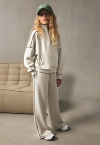 Beige sweatshirt with zip collar and side pockets, paired with matching loose-fitting trousers and white sneakers. Green cap adds contrast.