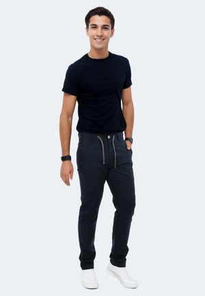 Schwarzes T-Shirt mit kurzen Ärmeln, kombiniert mit dunklen Marineblauen Hosen mit dekorativer Schnürung und Knopf vorne. Das Modell trägt weiße Turnschuhe.