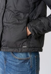 Giacca piumino nera con finitura testurizzata, tasca con zip e dettaglio logo argentato. Abbinata a jeans di denim blu con cuciture visibili.