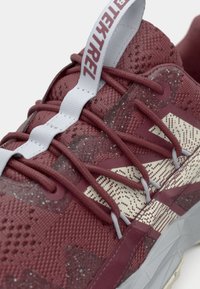 Primo piano di una sneaker color bordeaux con strisce bianche a motivo, lacci bordeaux e un'etichetta bianca con testo del marchio.