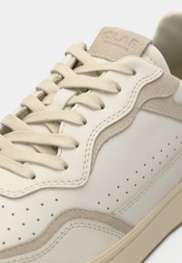 Close-up de um ténis de couro off-white com detalhes em camurça bege, perfurações ao longo do lado e atacadores planos bege passados por ilhós.