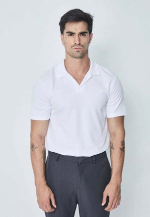 Homme aux cheveux courts et foncés, portant un polo blanc et un pantalon foncé, debout devant un fond clair uni, face à l'avant.