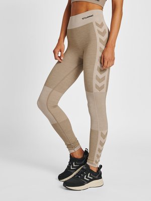 Persoon draagt beige Hummel legging met chevronpatronen aan de zijkanten en zwarte sportschoenen met witte zolen, staand tegen een effen achtergrond.
