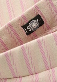 Gebreide stof met een beige en roze zigzagpatroon. Bevat een donker label en een transparante knoop. Zachte textuur met subtiele glans.