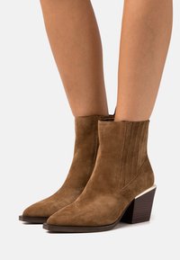 Stivaletti in suede marrone con punte affusolate, tacco a blocco impilato e dettagli cuciti verticalmente sui lati, indossati su gambe nude.