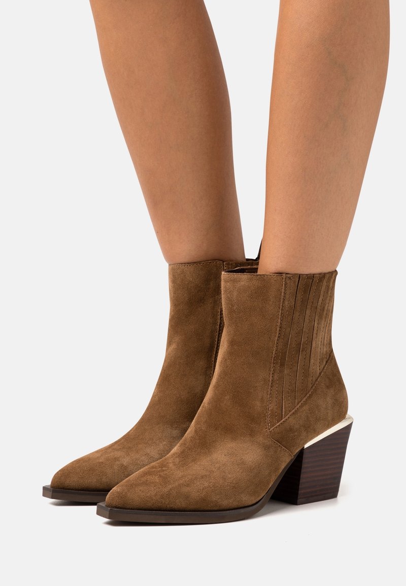 Stivaletti in suede marrone con punte affusolate, tacco a blocco impilato e dettagli cuciti verticalmente sui lati, indossati su gambe nude.