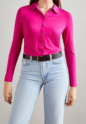 Nainen, jolla on kirkkaan vaaleanpunainen pitkähihainen napitettava paita, joka on pantu vaaleansinisten, korkeavyötäröisten farkkujen sisään, ja musta vyö.