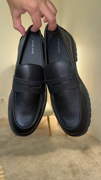 Main tenant une paire de mocassins en cuir noir sans lacets avec la marque "Pier One" à l'intérieur, sur un fond intérieur beige.