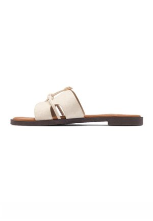 Sandalia plana beige con puntera abierta, suela oscura y tiras anchas cruzadas con pequeño nudo y detalle de hebilla en el lateral.