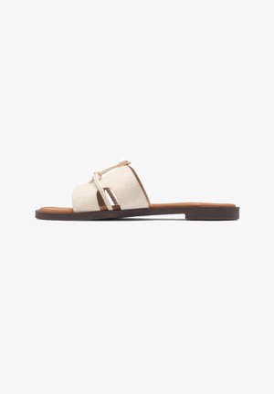 Flad beige åben-tå slide sandal med mørk sål og brede krydsede stropper med lille knude og spænde detalje på siden.