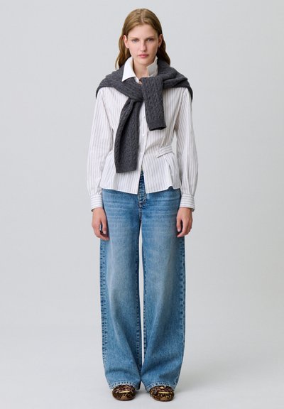 Camicia bianca a righe con vita aderente, abbinata a jeans azzurri con gamba ampia. Un maglione grigio a trecce è appoggiato sulle spalle.