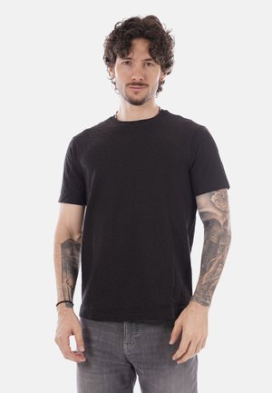 Mann mit lockigem Haar, der ein schwarzes T-Shirt und graue Jeans trägt, mit detaillierten Tätowierungen auf beiden Unterarmen, stehend vor einem einfarbigen Hintergrund.