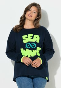 Frau trägt einen marineblauen Pullover mit neongrünem "SEA WAVE"-Schriftzug und Unendlichkeitssymbol in Form einer Welle, kombiniert mit blauen Jeans, steht vor einer weißen Wand.