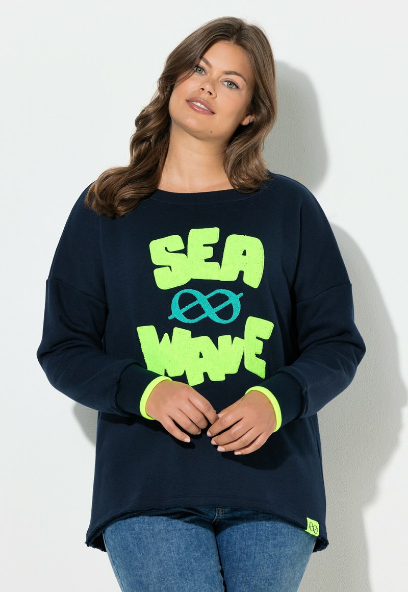 Frau trägt einen marineblauen Pullover mit neongrünem "SEA WAVE"-Schriftzug und Unendlichkeitssymbol in Form einer Welle, kombiniert mit blauen Jeans, steht vor einer weißen Wand.