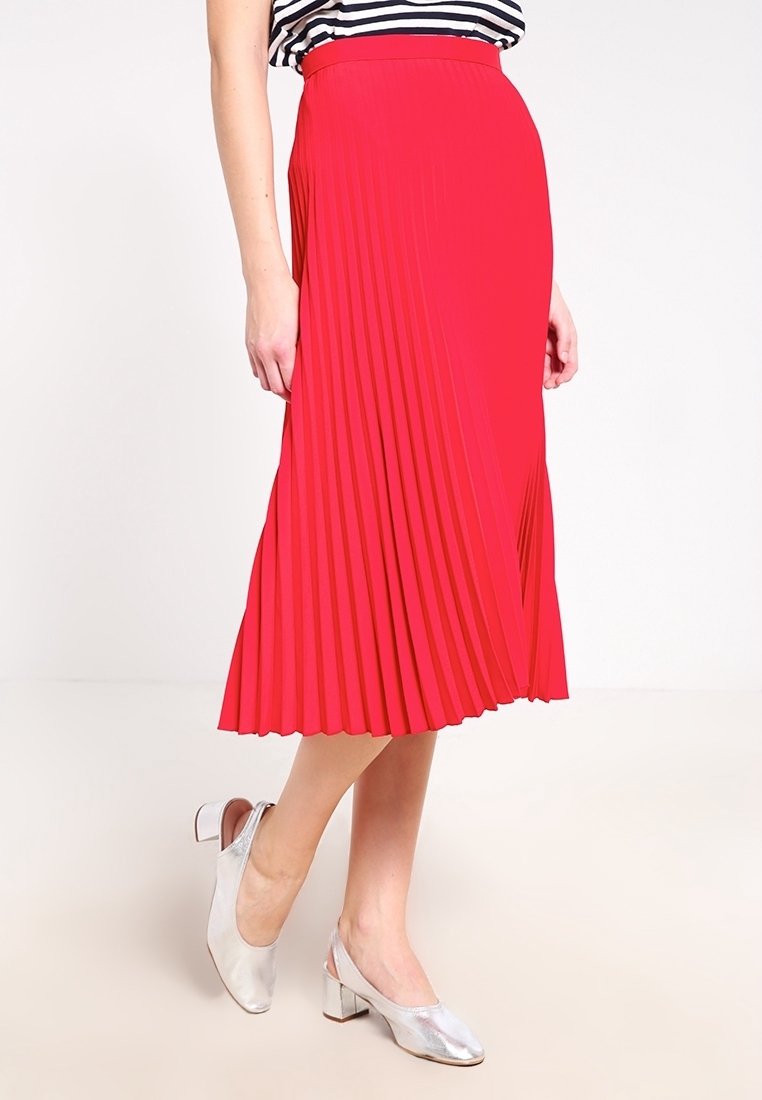 Falda midi plisada roja con cintura ajustada, hecha de tejido ligero; combinada con una camiseta de rayas y tacones plateados. La falda presenta pliegues verticales marcados.