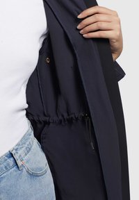 L'interno della giacca navy presenta un elastico con coulisse, una texture di tessuto liscia e un bottone in metallo dorato. Abbinata a una camicia bianca e denim.