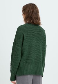 Mörkgrön stickad tröja med diamantmönster, ribbad textur och vida ärmar, visad bakifrån. Långa ärmar och avslappnad passform.