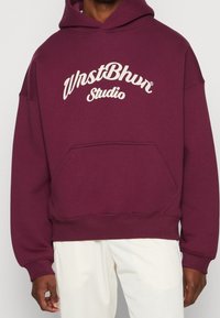 Man med en vinröd huvtröja med vit text "WnstBhm Studio", i kombination med vita byxor som visar framfickorna och långa ärmar.