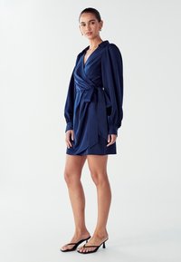 Námorníckomodrá zavinovacia šaty s dlhými nafúknutými rukávmi a opaskom na páse, vyrobené z hladkej, lesklej látky a s krátkym lemom.