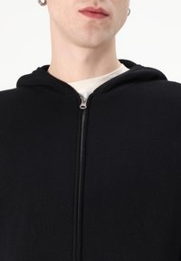 Svart zip-up hoodie i ribbat tyg, med en huva som har dragsko och en silverfärgad dragkedja. Det inre lagret är en ljusfärgad tröja.