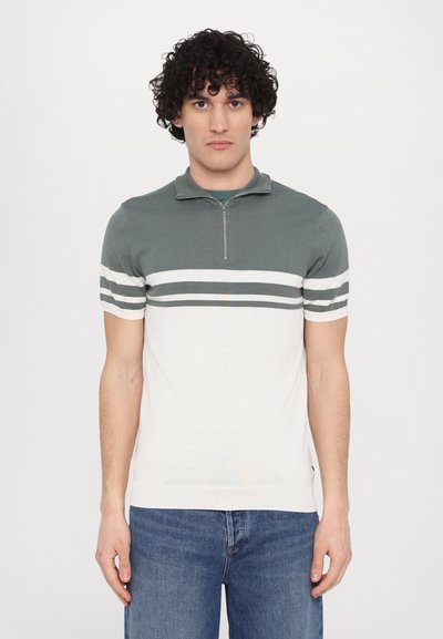 Tricou polo cu mânecă scurtă și guler cu fermoar, având o secțiune superioară teal și o secțiune inferioară albă cu dungi verzi închise. Textură din amestec de bumbac.