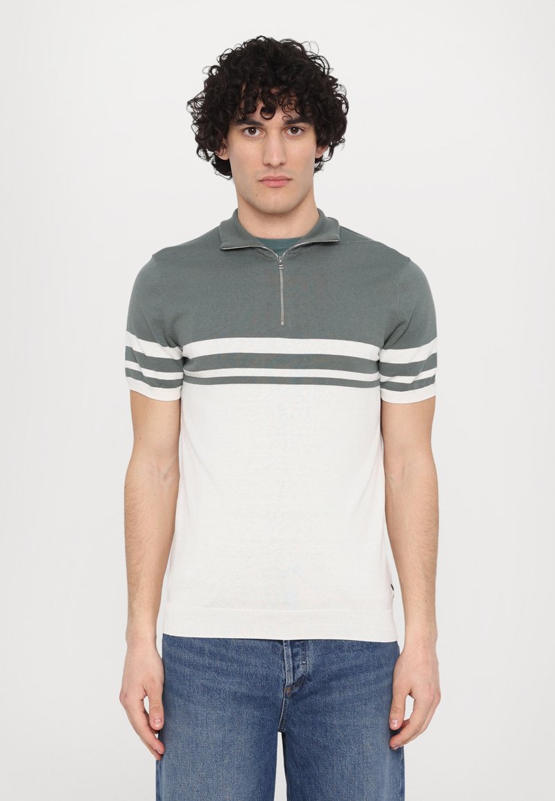 Kortärmad polo med dragkedja i kragen, med en teal övre del och en vit nedre del med mörkgröna ränder. Blandning av bomull.