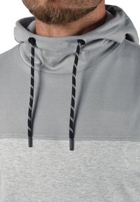Lys grå hettegenser med høy krage, med snor og svart-hvite mønstre. Glatt tekstur, minimalistisk design og ingen synlige logoer.