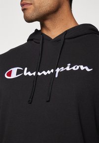 Sweatshirt à capuche noir avec un logo "Champion" brodé en blanc et un accent rouge. Le matériau semble doux avec des cordons de serrage au niveau du cou.