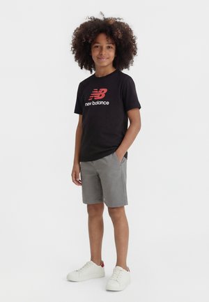 Barn med krøllet hår stående, iført sort New Balance t-shirt, grå shorts og hvide sneakers med røde detaljer.