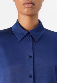 Chemise bleu marine à la texture lisse, dotée d'un col classique et de cinq boutons sur le devant, mettant en avant un design ajusté.